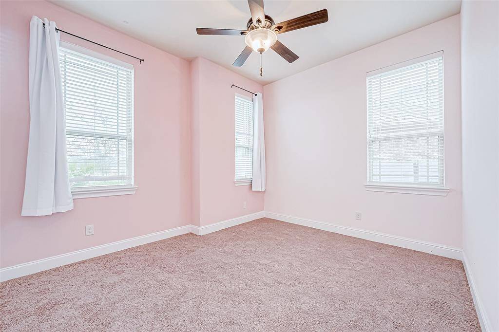 This room features soft pink walls, carpeted flooring, a ceiling fan with light, and two large windows with white curtains, offering a bright and cozy atmosphere.