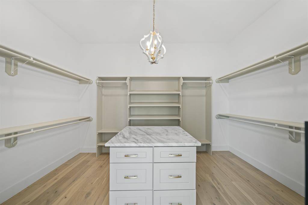 Spacious walk-in closet with hanging space, shelving, and a central island with drawers. Features a stylish chandelier and hardwood flooring. Ideal for organized storage.