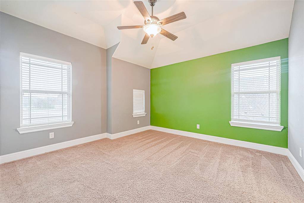 This bright room features a vaulted ceiling with a ceiling fan, neutral carpeting, and a striking green accent wall. It includes three windows that provide ample natural light, offering a fresh and inviting space.