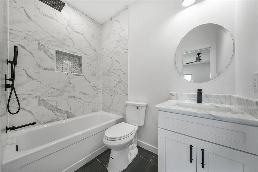 Attached bathroom features elegant marble tile walls, a sleek black shower fixture, a built-in shelf, a stylish vanity with a round mirror, and contemporary finishes.