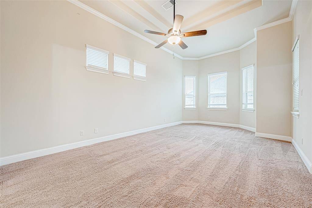 This spacious bedroom features neutral walls, a cozy carpet, and a tray ceiling with a ceiling fan. The room is brightened by multiple windows, including a charming bay window, perfect for natural light.