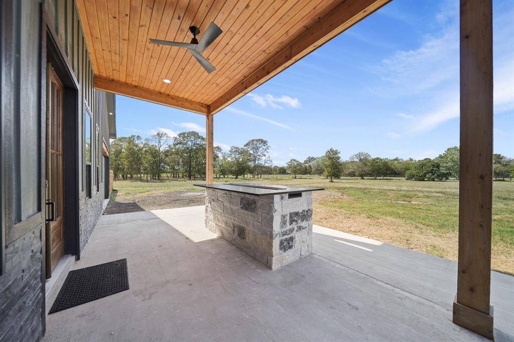 The back covered patio offers a stone bar height countertop with gas fire inset. It overlooks a spacious, open grassy area with trees in the distance, offering a serene outdoor setting.
