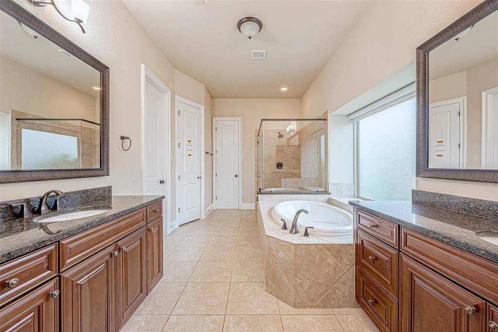 This spacious bathroom features dual sinks with granite countertops, a large soaking tub, a separate glass-enclosed shower, and elegant wooden cabinetry. Abundant natural light streams in through a frosted window, enhancing the room's bright, welcoming atmosphere.