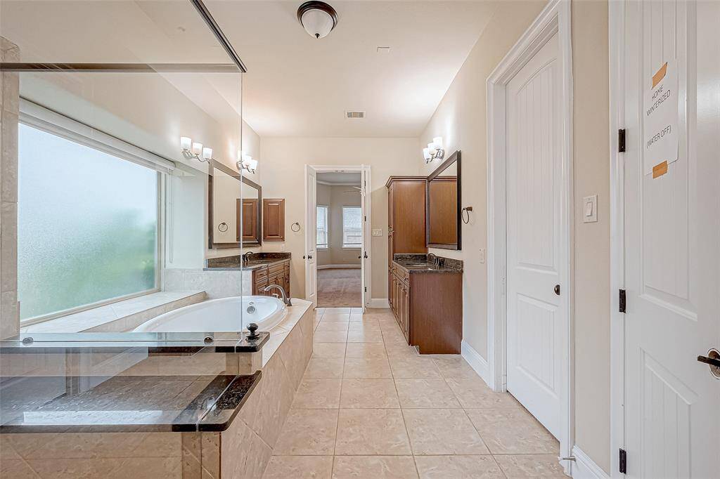 This spacious bathroom features a luxurious soaking tub with a large frosted window, dual vanities with ample storage, and elegant tile flooring. It leads into a well-lit adjoining room, adding to its convenience and appeal.