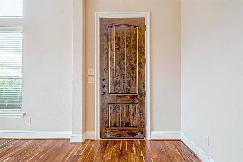 Showcases a beautifully crafted wooden door in rich tones, set against light walls. The room features elegant hardwood flooring and a window with white blinds nearby.