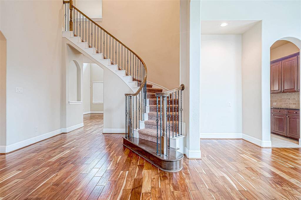 Spacious entryway with a gracefully curved staircase and wrought iron railings. The hardwood floors bring warmth, while open arches lead to a bright kitchen featuring dark wood cabinetry. High ceilings further elevate the airy, elegant atmosphere of the space.