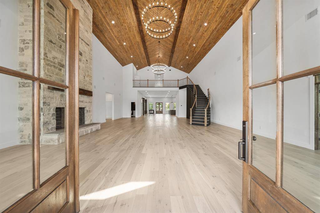 Open-concept living area features high vaulted wooden ceilings, a grand stone fireplace, and elegant chandeliers. The room is enhanced by natural light from large doors, with a stylish staircase leading to the second story. Perfect for modern living and entertaining.