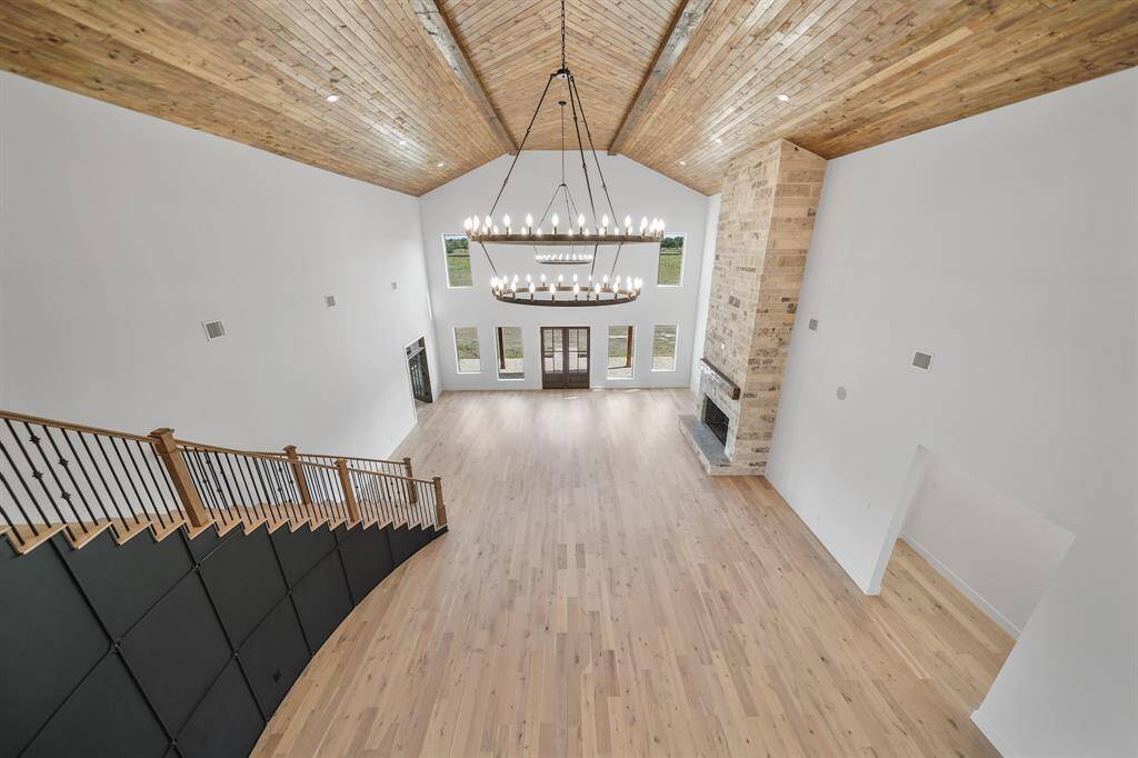 This spacious living area features a high vaulted 26ft wooden ceiling, elegant chandelier, and a grand staircase. It boasts a large stone fireplace and abundant natural light from tall windows, offering a warm and inviting atmosphere.