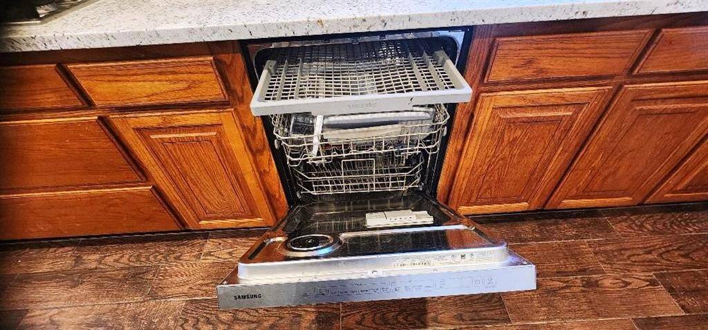 This kitchen features a built-in Samsung dishwasher with a sleek stainless steel finish, set against warm wooden cabinetry and a granite countertop. The dishwasher is open, revealing a spacious interior with multiple racks for efficient dishwashing.