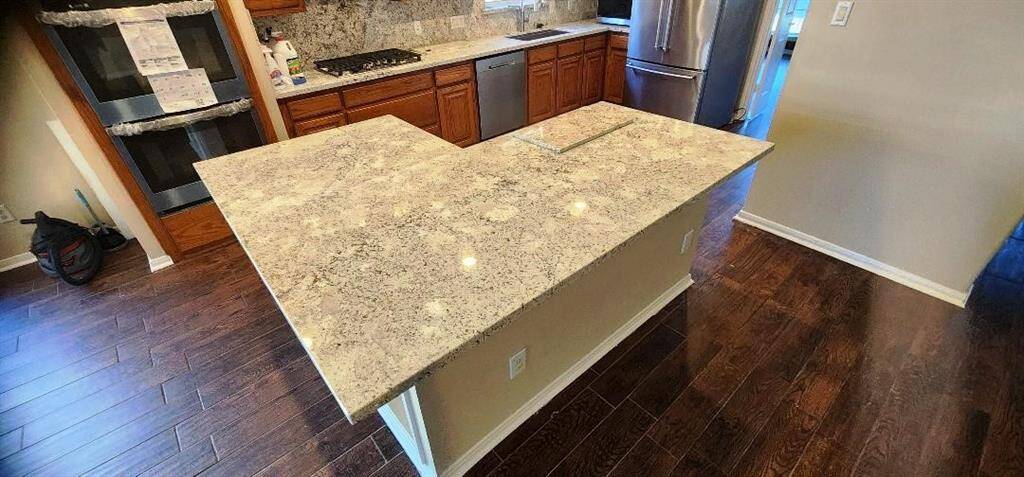 This kitchen features a large L-shaped granite countertop island, stainless steel appliances, wooden cabinetry, and dark hardwood flooring, offering a modern and spacious cooking area.