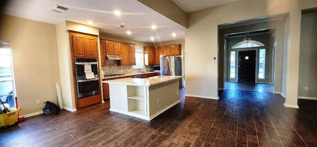 This photo showcases a spacious, open-concept kitchen and living area with dark wood flooring. The kitchen features wooden cabinets, stainless steel appliances, and a central island with a countertop. The area is well-lit with recessed lighting, and there's a view into a hallway leading to the entrance, which has large windows and a decorative light fixture.