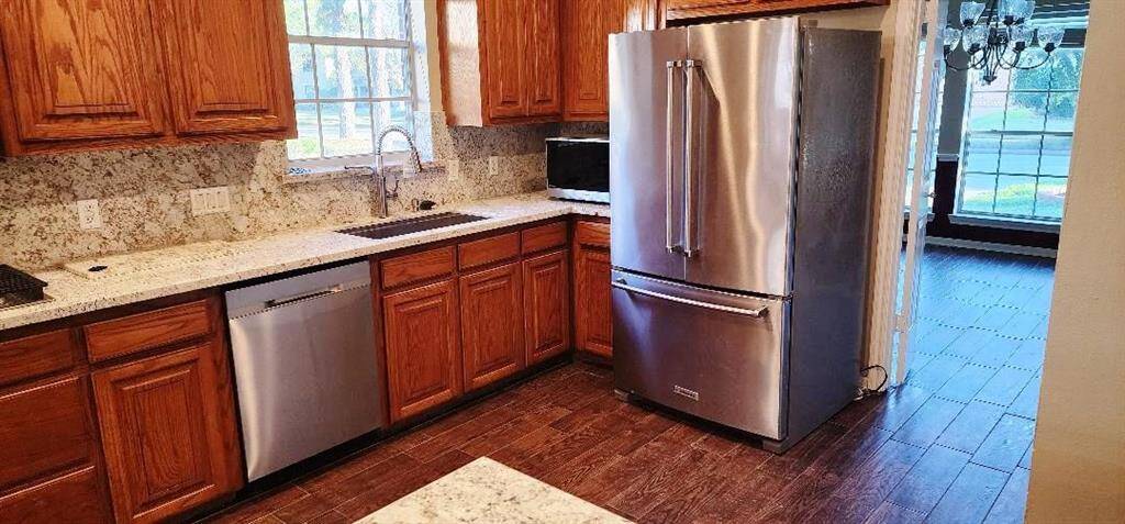 This kitchen features warm wood cabinetry, granite countertops, and stainless steel appliances, including a French-door refrigerator. The space is well-lit with a large window over the sink and opens to a dining area with a view outside.