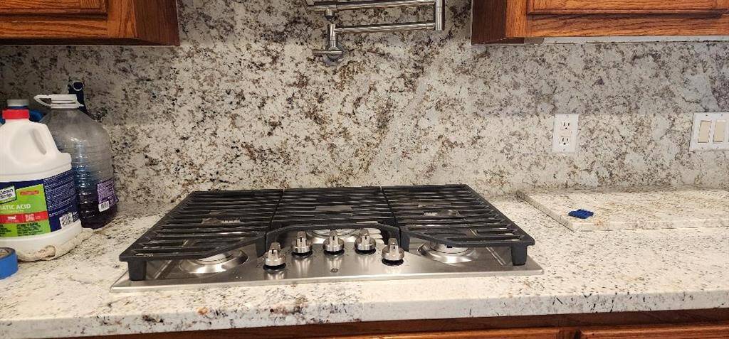 This kitchen features a sleek gas cooktop set in a granite countertop with matching backsplash. It includes wooden cabinetry above, a pot filler faucet for convenience, and a modern, clean look.