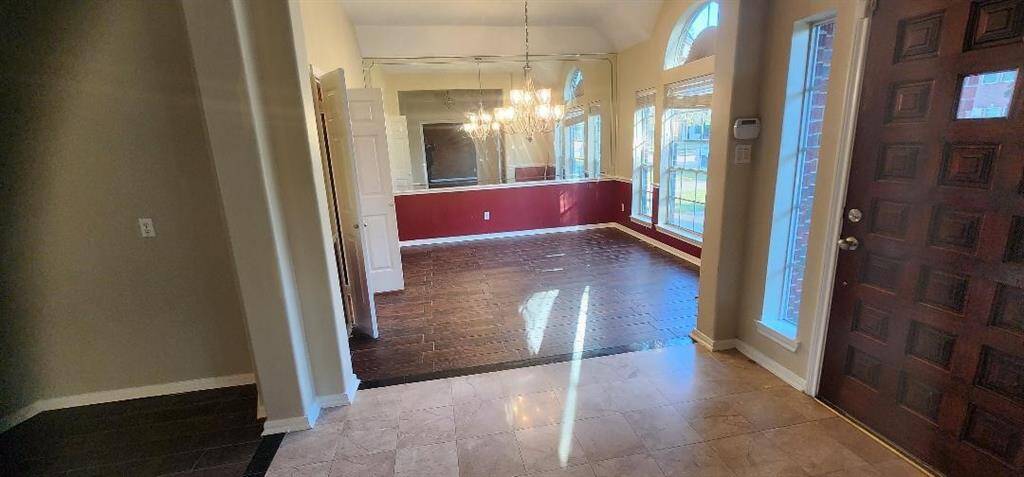 This photo showcases a welcoming entryway with a rich wooden front door and tiled flooring that transitions into a dining area. The dining space features dark wood flooring, a chandelier, and large windows that provide ample natural light. The high ceiling and open layout create an inviting and spacious atmosphere.