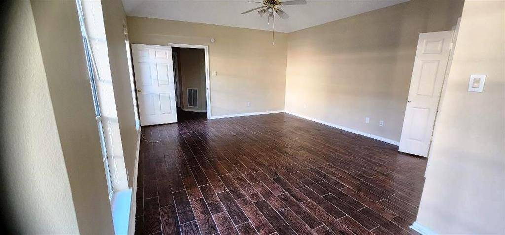 This room features dark wood flooring, neutral walls, and ample natural light from large windows. It has a ceiling fan and two doors, offering a spacious and versatile living area.