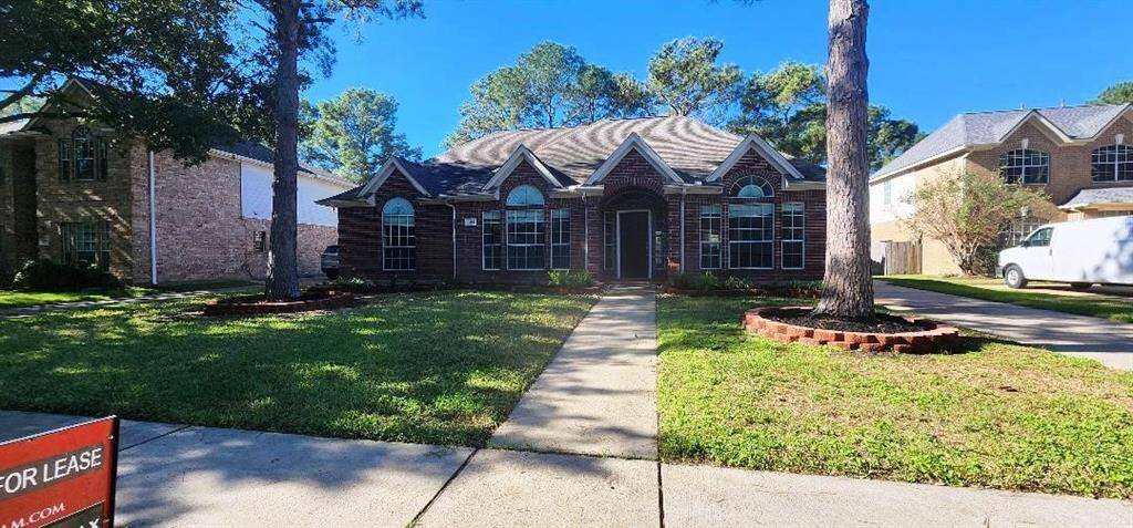 This photo shows a charming single-story brick home with a manicured lawn and mature trees. It features large arched windows and a central pathway leading to the entrance. The property includes a driveway and is located in a suburban neighborhood.