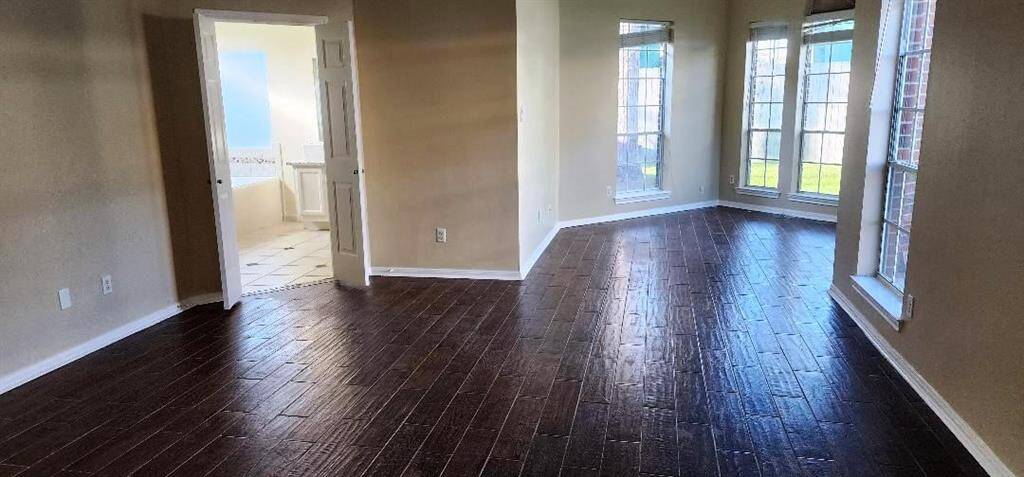 Spacious room with dark wood flooring and ample natural light from large windows. Neutral walls and an open doorway leading to a bright adjacent room with tile flooring. Ideal for versatile living or dining space.