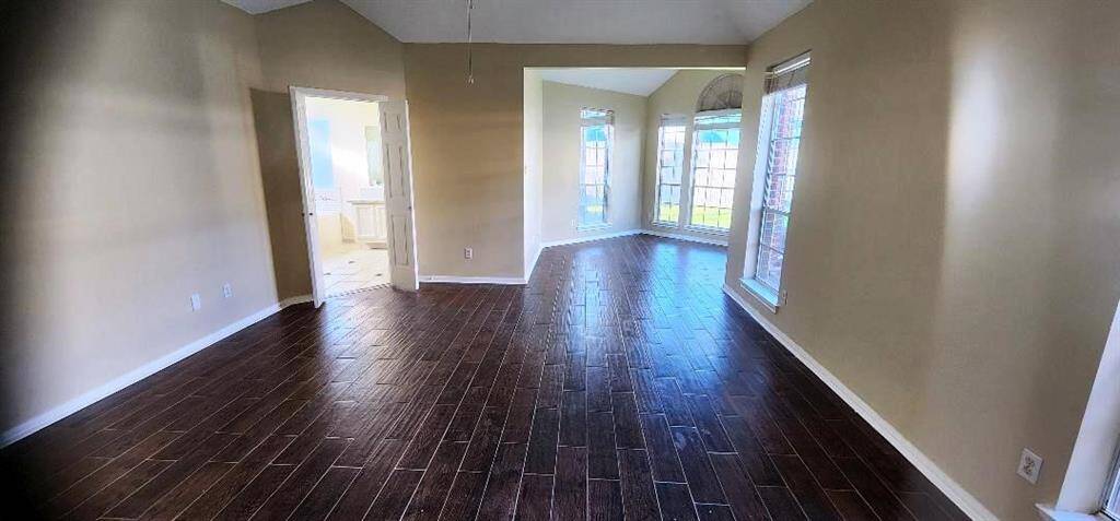 This photo showcases a bright, open living space with dark wood flooring and neutral walls. The room features large windows that provide ample natural light. There's an adjoining room visible through a doorway, enhancing the open feel of the layout.