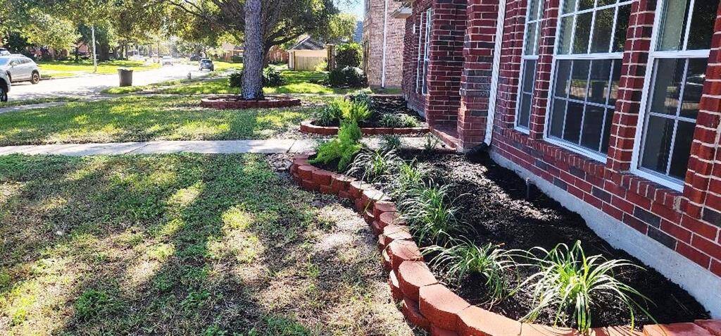 The photo showcases the front yard of a home with a well-maintained lawn and a brick house. There's a tidy flower bed with new plants along the house, bordered by decorative brickwork. A tree provides shade, and the neighborhood appears quiet and residential.