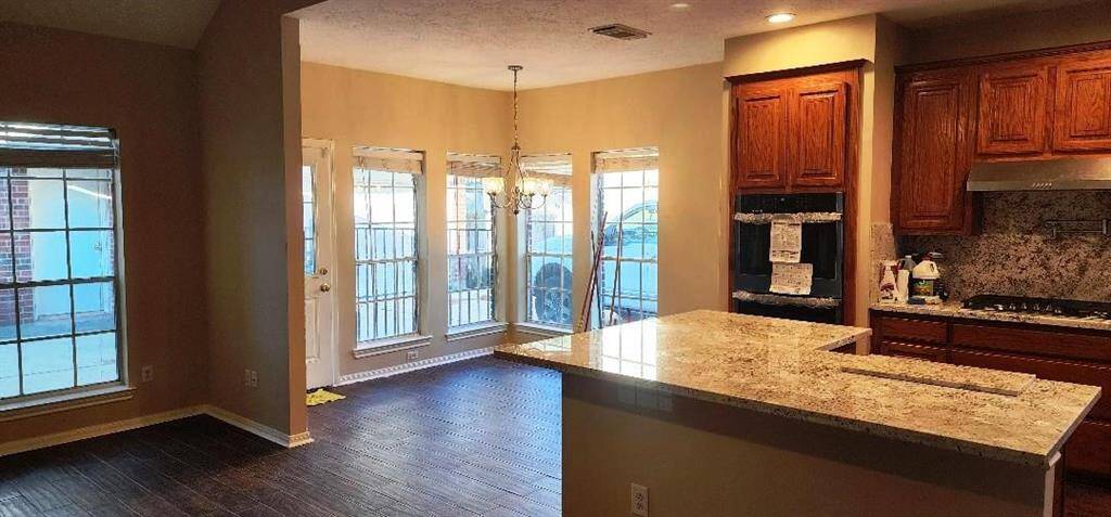 This photo shows a bright, open-concept kitchen and dining area. The space features a large granite countertop island, wooden cabinets, a built-in oven, and a gas stovetop. The dining area is enhanced by multiple windows and a glass door, allowing for plenty of natural light. Warm-toned walls and dark flooring create a cozy atmosphere.