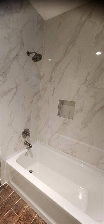 This bathroom features a modern bathtub with elegant marble-style wall tiles and a built-in niche for toiletries. The showerhead and faucet have a sleek, contemporary design, complemented by warm-toned wood-look flooring.