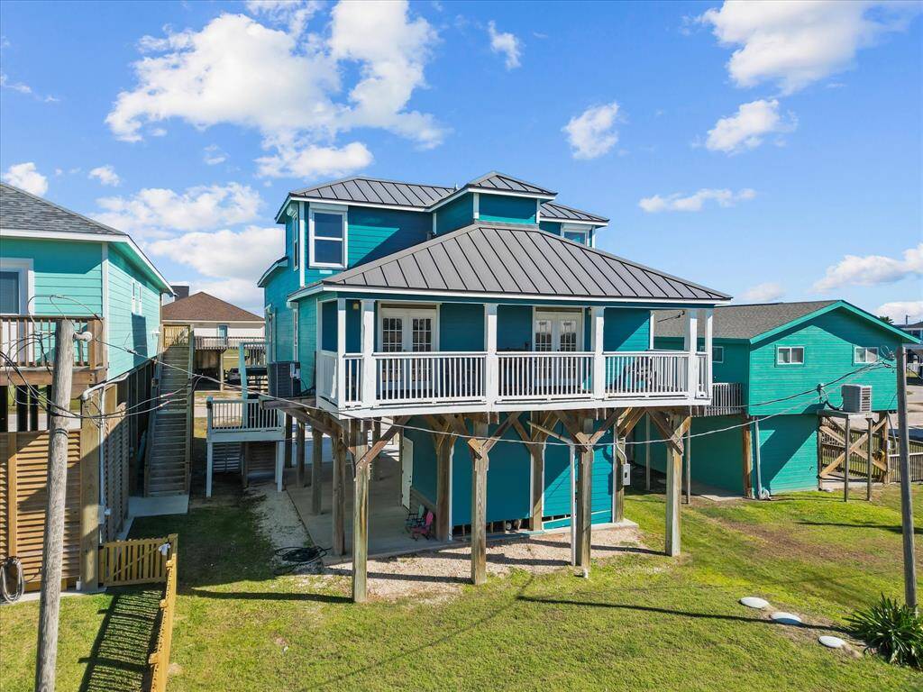 Elevated coastal home with a bright blue exterior, spacious driveway, and a welcoming front porch. Features include stilts for flood protection and open under-house parking. Ideal for beachside living.