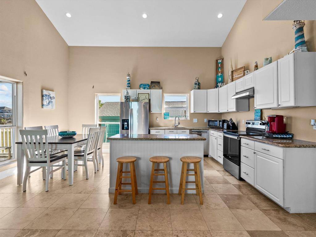 This spacious kitchen features high ceilings, modern stainless steel appliances, and ample white cabinetry. It includes a central island with barstools and an adjoining dining area with natural light, offering a bright and welcoming atmosphere.