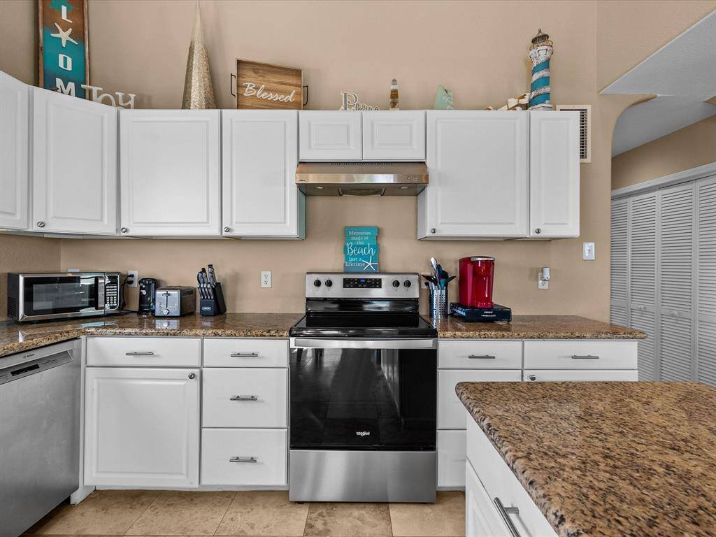 This kitchen features white cabinets, granite countertops, and stainless steel appliances, including a modern stove and dishwasher. It has a cozy, beach-themed decor with ample storage and counter space for cooking.