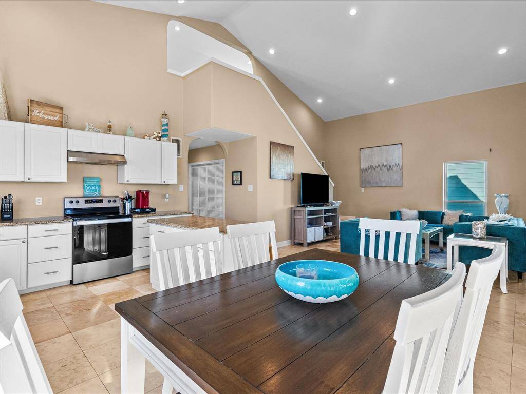 This open-concept living space features high ceilings and a cozy seating area. The modern kitchen boasts stainless steel appliances and ample white cabinetry, while the dining area offers a warm, inviting atmosphere with a large wooden table. Natural light enhances the bright, airy feel of the room.