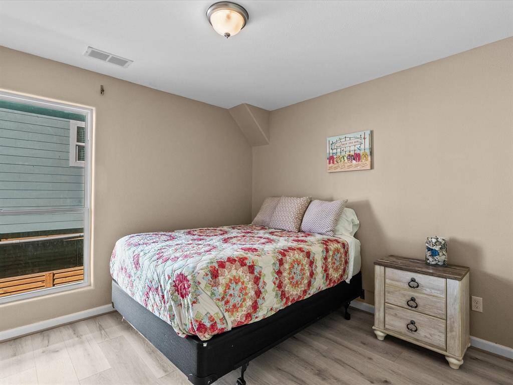 Cozy bedroom with a large window providing natural light, neutral walls, and wood-style flooring. Features a comfortable bed with colorful bedding and a small nightstand with drawers.