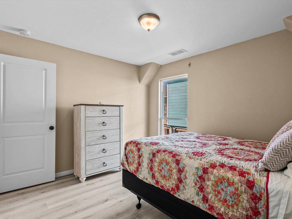 Cozy bedroom with neutral walls and laminate flooring. Features a window for natural light, a white door, and a five-drawer chest. The bed has a colorful quilt, creating a warm, inviting atmosphere.