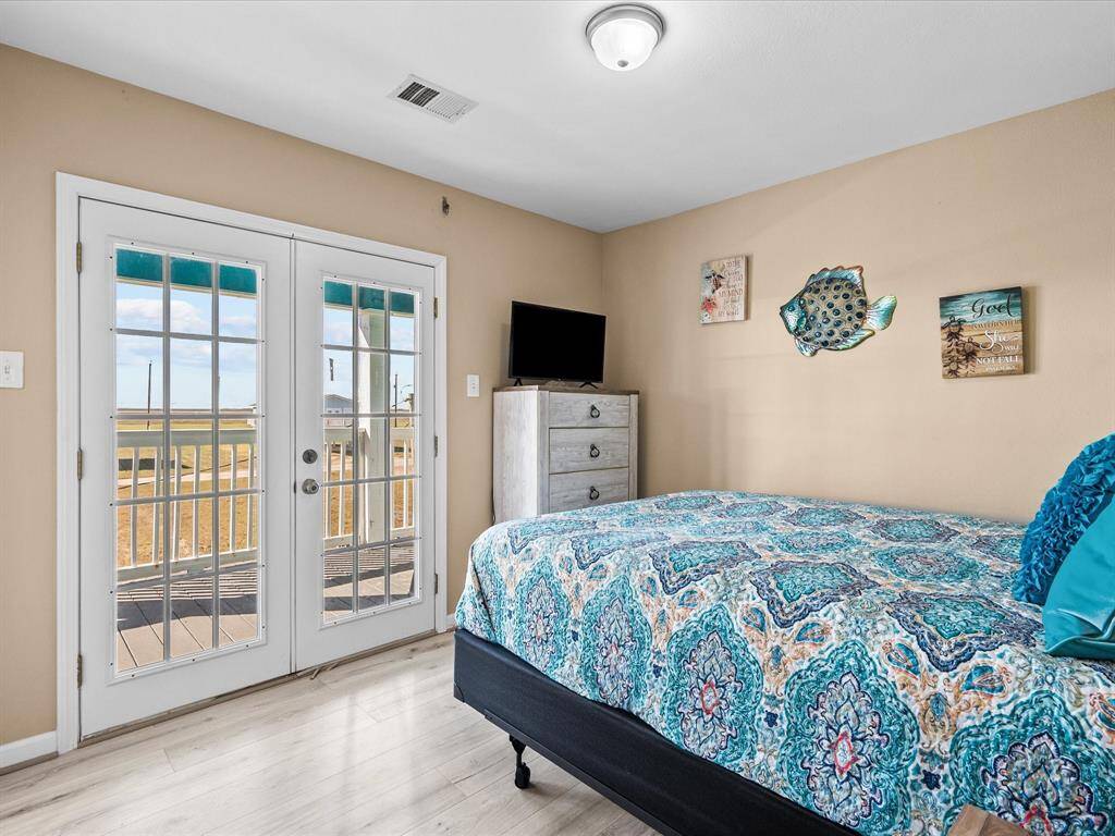 This cozy bedroom features a large bed with a colorful patterned comforter, light wood flooring, and beige walls. French doors open to a balcony with scenic views, enhancing natural light. Wall decor adds charm, and there's a small dresser with a TV.