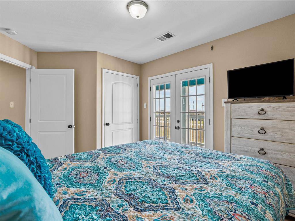 Cozy bedroom with French doors leading to a balcony, offering natural light. Features a decorative bedspread, ceiling light, and a dresser with a mounted TV. Neutral walls and white doors create a clean, inviting space.