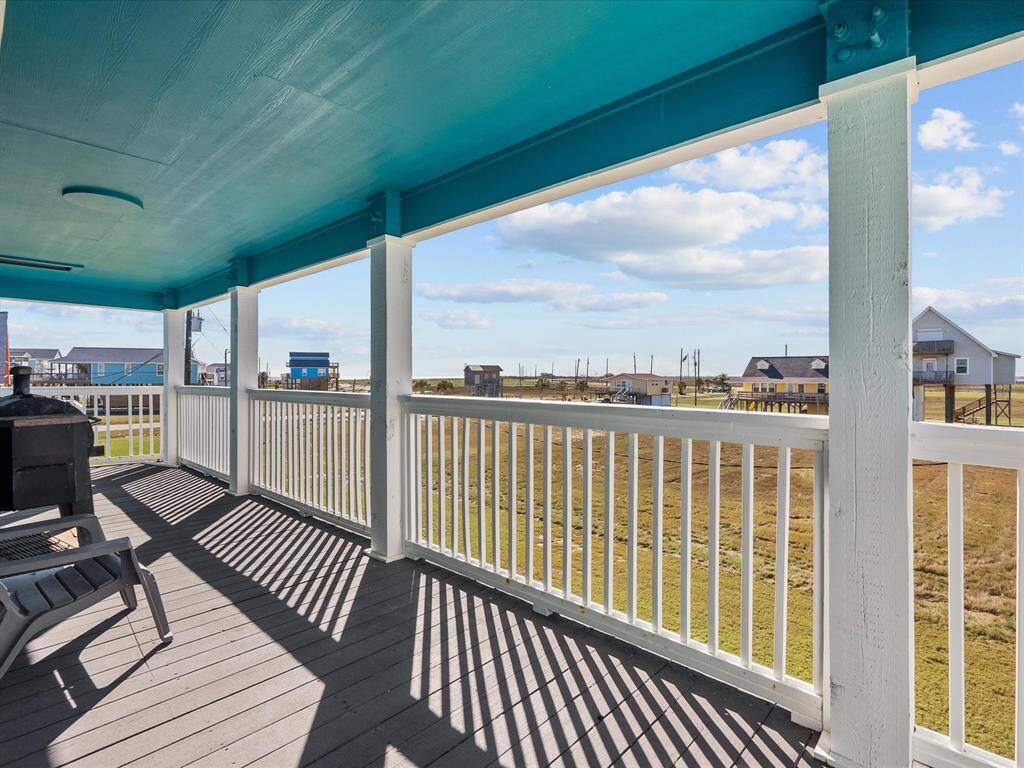 This photo showcases a covered outdoor deck with a bright blue ceiling, offering ample space for relaxation. It features white railings and provides a scenic view of a grassy area and nearby houses, perfect for enjoying the outdoors.