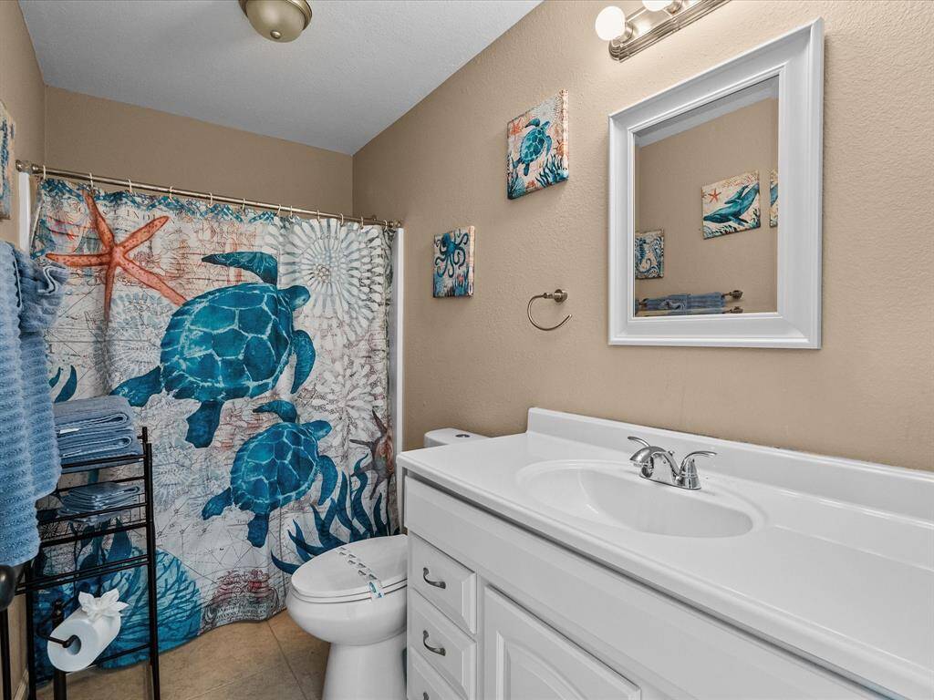 This bathroom features a nautical theme with a turtle shower curtain and matching decor. It includes a white vanity with a single sink, a wall-mounted mirror, and soft beige walls. The space is complemented by blue towels and a small storage rack.