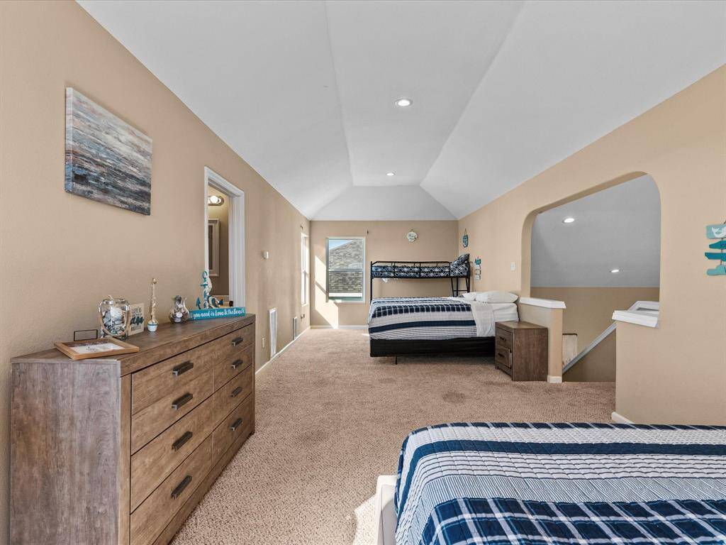 This spacious, carpeted bedroom features two single beds and a bunk bed, with soft beige walls and recessed lighting. A large dresser offers ample storage, and the room has a cozy, inviting atmosphere. An archway leads to the staircase.