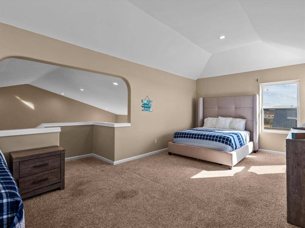 This spacious bedroom features a cozy carpet, a large bed, and ample natural light from a window. The room has an open layout with a partial wall divider and recessed lighting, creating a bright and inviting atmosphere.