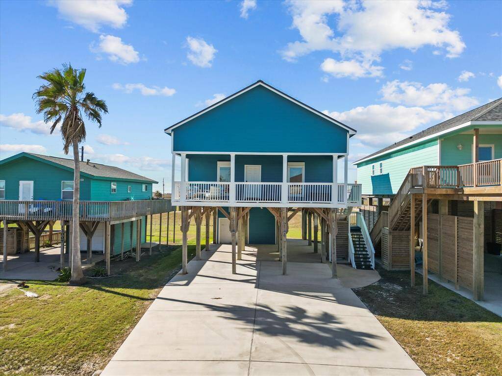 This elevated beach house features a vibrant blue exterior with a spacious driveway and covered parking. It includes a front porch with white railings, set against a backdrop of palm trees and clear skies. Perfect for coastal living.