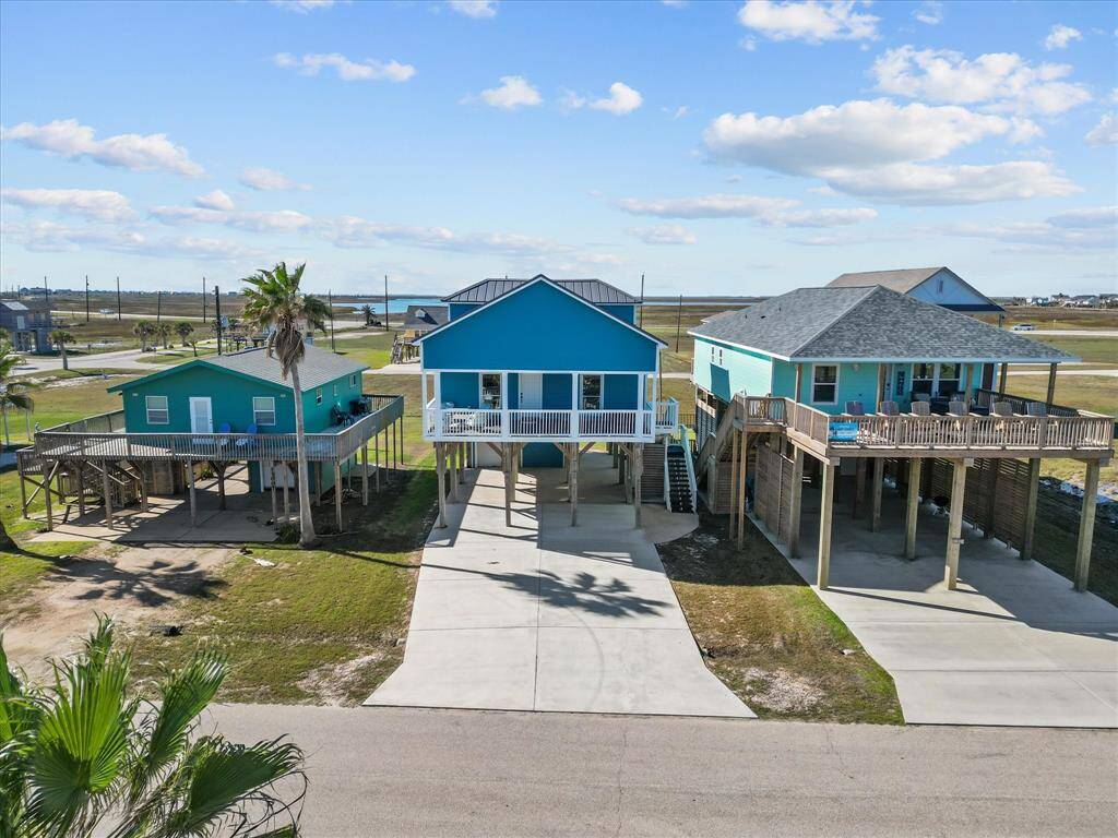 This photo shows three elevated beach houses, each with vibrant blue exteriors. They feature spacious decks and are set on stilts with driveways underneath. The surrounding area is open and coastal, offering views of nearby water and flat terrain.
