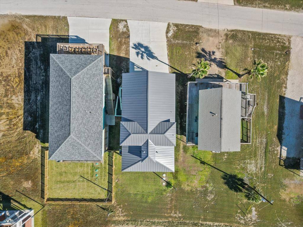 This aerial view shows three adjacent homes with well-maintained lawns and driveways. The central house features a modern metal roof, while the neighboring houses have traditional shingled roofs. There's a clear street view and palm trees in the yards, indicating a suburban or coastal setting.