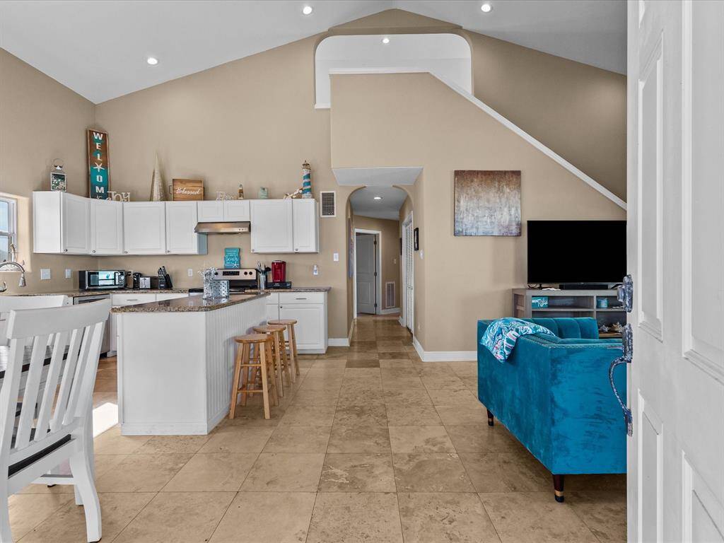 This open-concept living area features high ceilings and natural light. The kitchen has white cabinets, granite countertops, and a central island with bar seating. The space flows into a cozy living area with a vibrant blue sofa and a large TV, perfect for modern living.