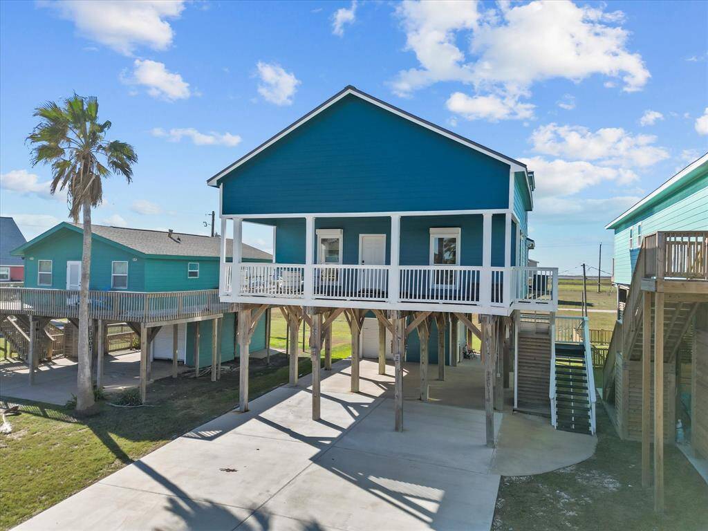 This elevated coastal home features vibrant turquoise siding with white trim, a spacious front porch, and a covered parking area beneath. Ideal for beachside living with scenic views.