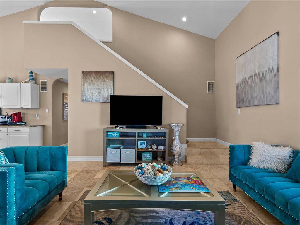 Bright and airy living room with vaulted ceilings, featuring modern decor and teal accents. The space includes a flat-screen TV, two plush sofas, and a glass coffee table, offering a cozy yet stylish ambiance. A staircase leads to an upper level, and the open layout flows into the kitchen area.