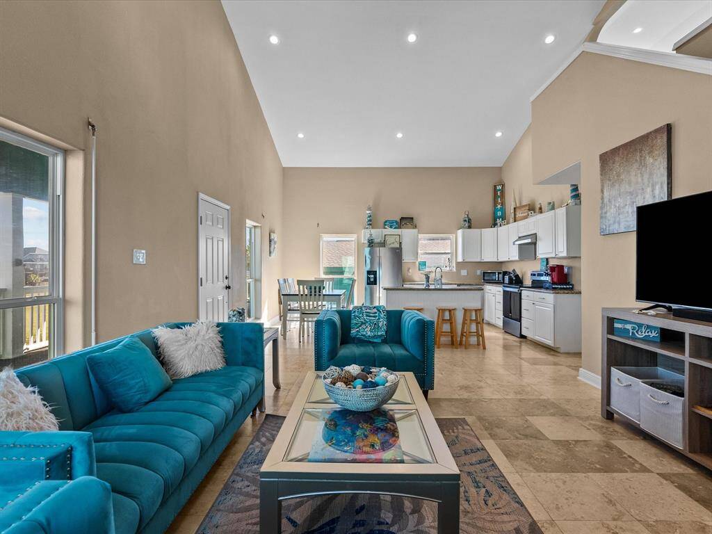 This spacious open-concept living area features high ceilings, abundant natural light, and modern finishes. It includes a stylish kitchen with white cabinetry, a cozy sitting area with teal sofas, and a dining space, all seamlessly integrated for comfortable living.