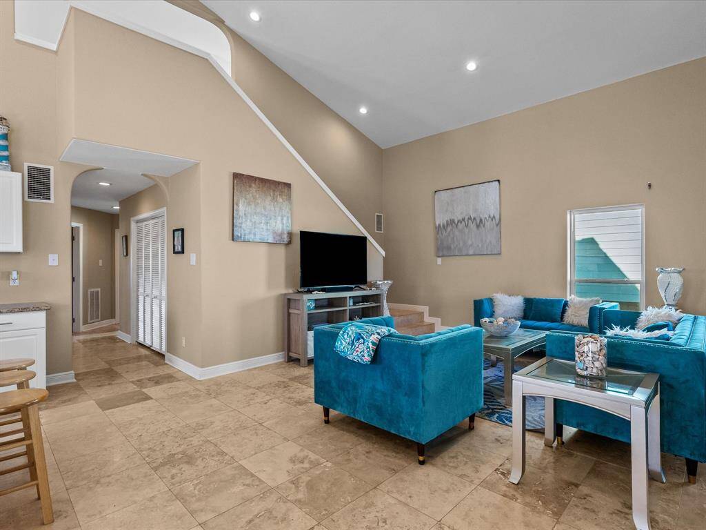 This spacious living room features high ceilings and elegant tile flooring. It's well-lit with recessed lighting and has a modern, cozy feel with teal velvet sofas. The room includes a TV setup and leads to a hallway with a closet, offering a welcoming and stylish space for relaxation and entertainment.