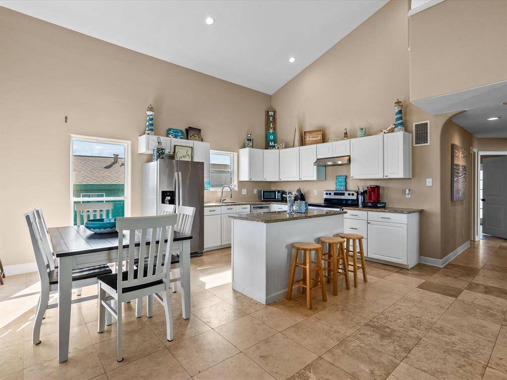 Bright, open kitchen with vaulted ceilings, featuring modern stainless steel appliances, white cabinetry, and an island with bar stools. Adjacent dining area with a table and chairs, tile flooring throughout, and ample natural light.