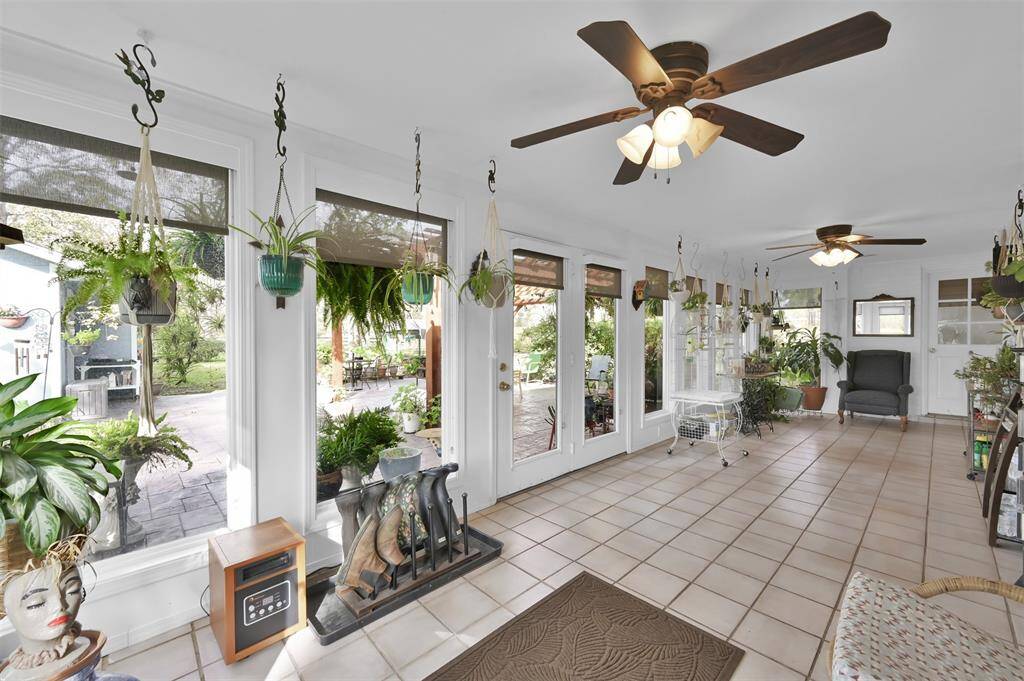 Bright sunroom with large windows, ceiling fans, and tiled floor. It features numerous hanging plants and overlooks a lush garden, providing a serene indoor-outdoor feel