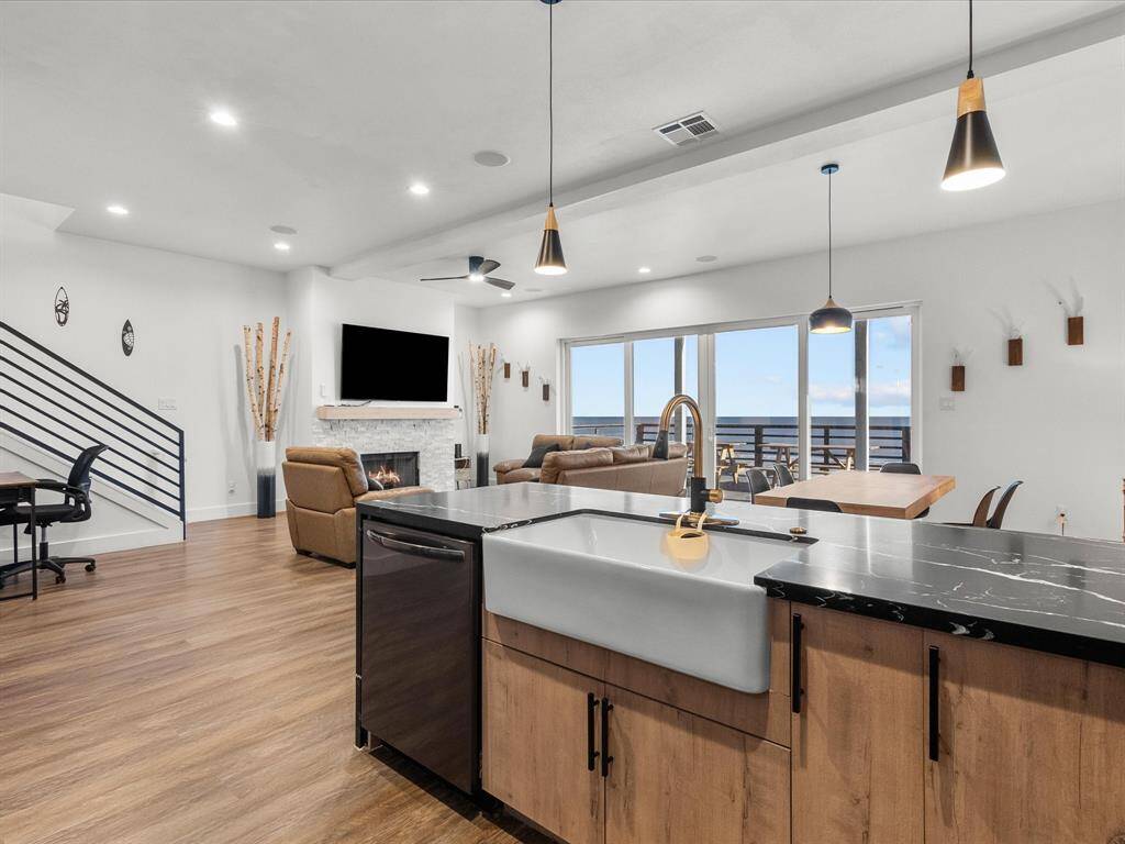 This open-concept living space features a modern kitchen with a farmhouse sink and sleek pendant lighting. It flows into a cozy living area with a fireplace and large windows offering ocean views. The room is bright with wood flooring and contemporary decor, providing a spacious and inviting atmosphere.