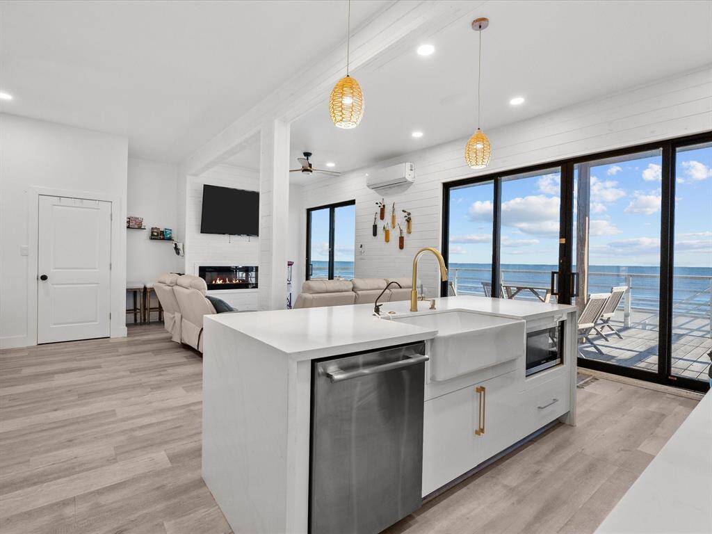 This modern, open-concept living space features a bright kitchen with a large island, sleek appliances, and a farmhouse sink. It opens to a cozy living area with a fireplace and large sliding glass doors that lead to a deck with stunning ocean views.