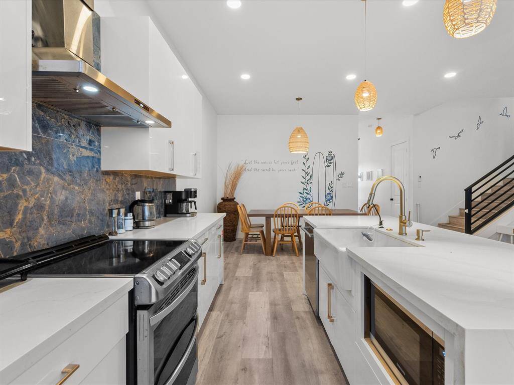 Modern kitchen and dining area with sleek white cabinetry, marble backsplash, and stainless steel appliances. Bright and inviting with hanging pendant lights, wood flooring, and a cozy dining nook. Artistic wall accents add a stylish touch.